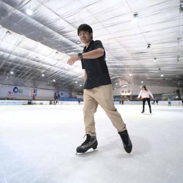 A man in black shirt is skating on ice rink with smiling face while the woman is skating behind